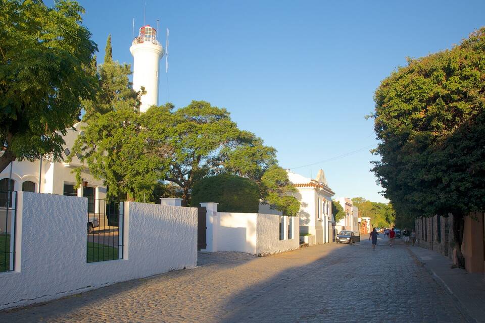 Una strada acciottolata fiancheggiata da edifici dalle pareti bianche, un faro e alberi di verde intenso.