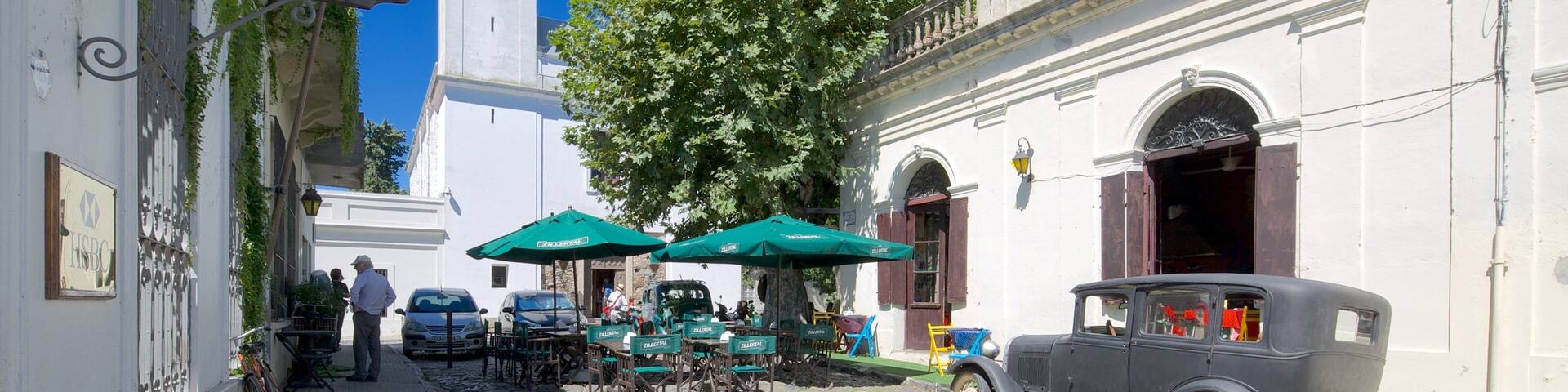 A cobblestone street with vintage cars, outdoor seating, and a clock tower.