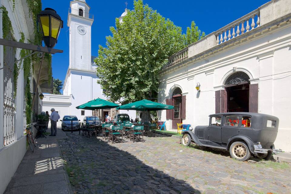 Una strada acciottolata con auto d'epoca, posti a sedere all'aperto e una torre dell'orologio.