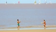 Two people playing with frisbees on a beach with sailboats in the background.