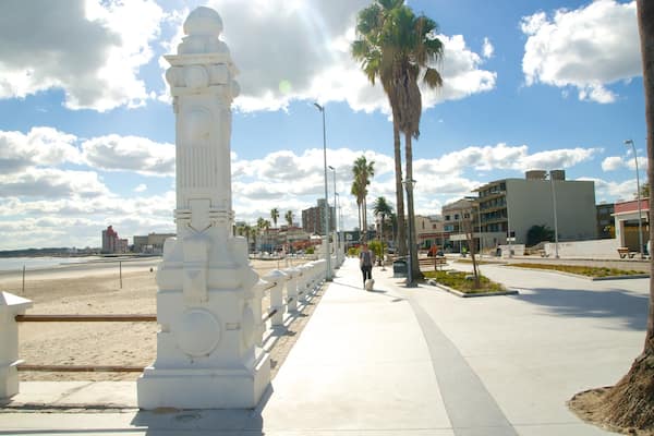 En kystpromenade med et hvidt monument, palmer og moderne bygninger.