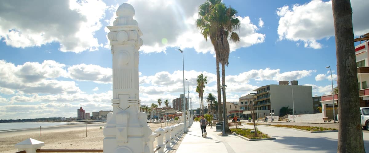 A coastal promenade with a white monument, palm trees, and modern buildings.
