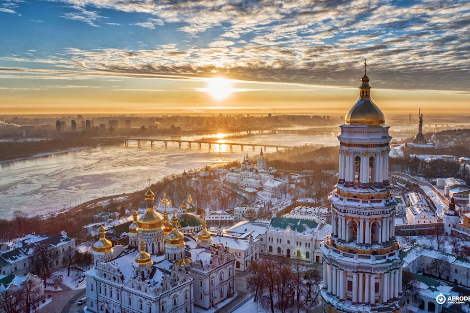 A winter cityscape with a prominent church tower, golden domes, and a frozen river under a sunset sky.
