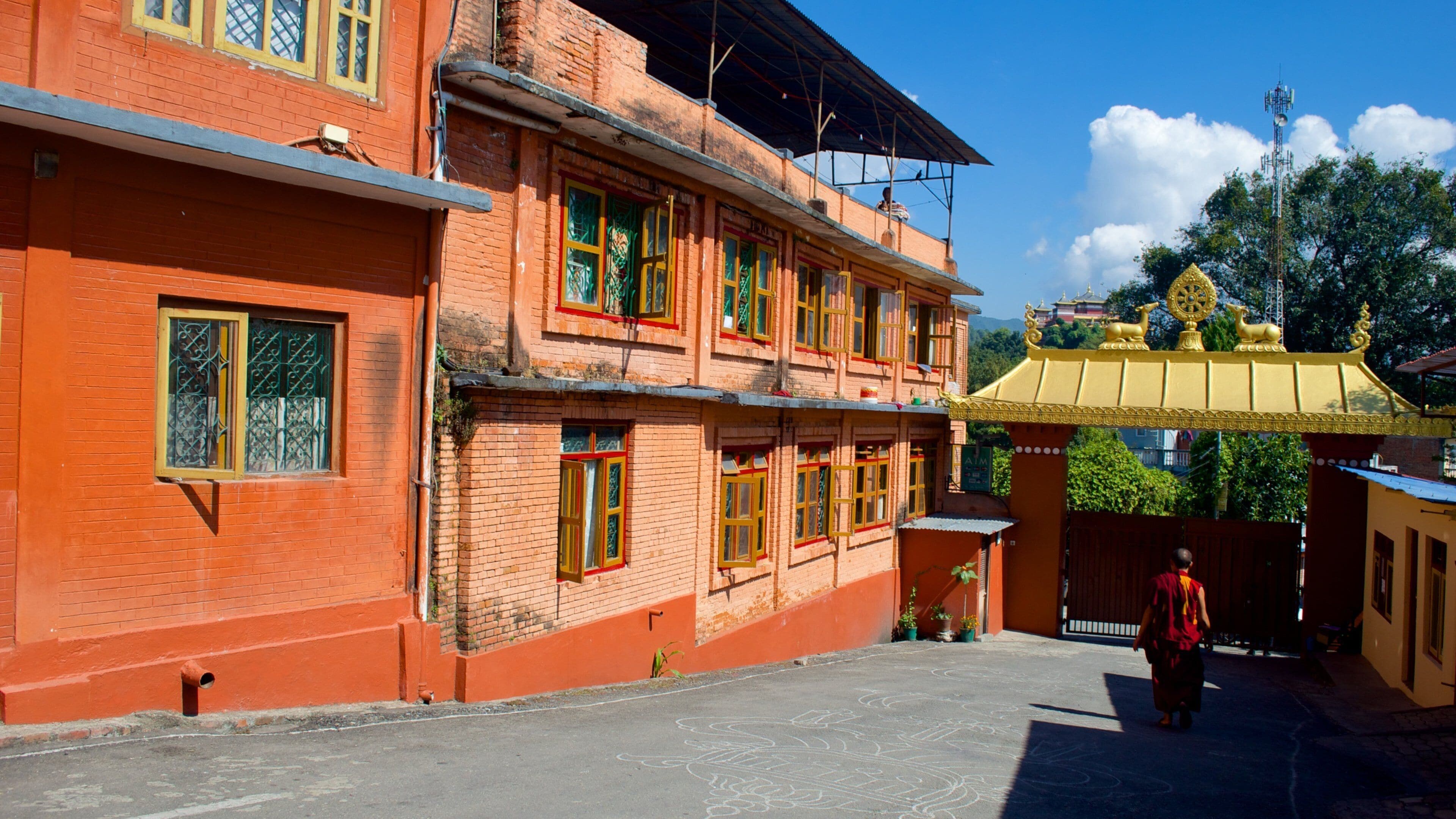 A traditional building with red brick walls and yellow roofs, a person walking in front, and a temple with a golden roof in the background.