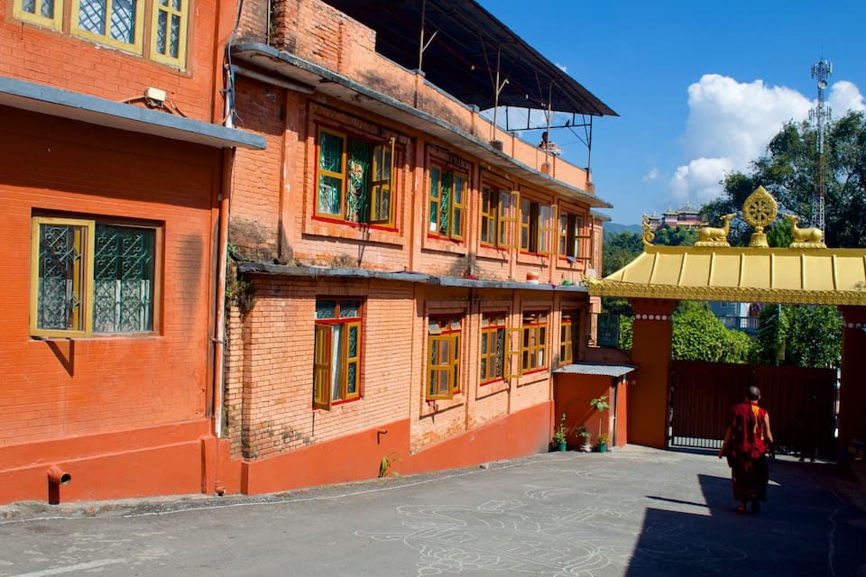 A traditional building with red brick walls and yellow roofs, a person walking in front, and a temple with a golden roof in the background.