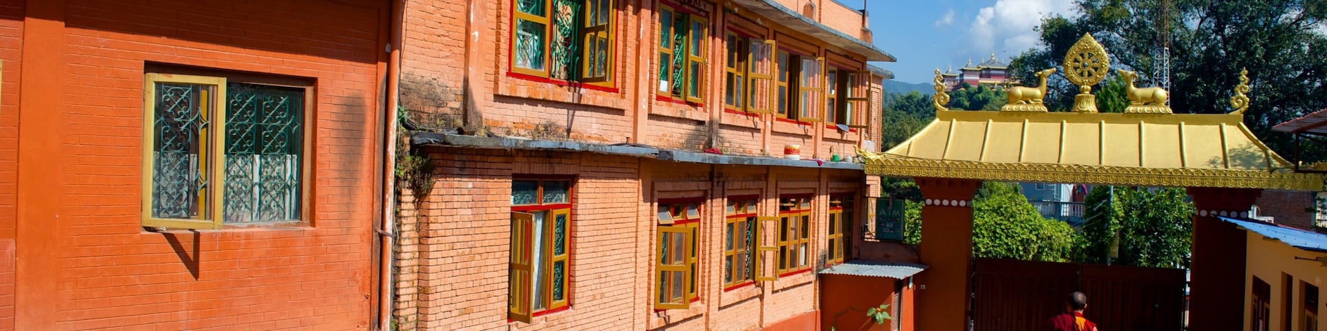 Un edificio tradicional con paredes de ladrillo rojo y techos amarillos, una persona caminando al frente y un templo con techo dorado al fondo.