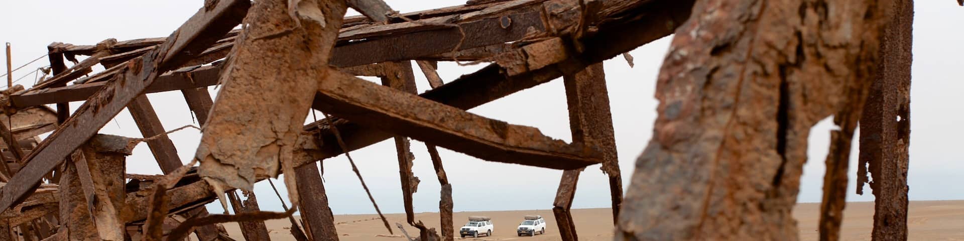 A rusted metal structure in a sandy desert with two white vehicles in the background.