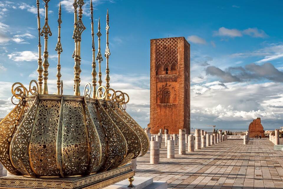 A large ornate metal structure with intricate designs, a tall brick tower with a decorative facade, and a paved pathway leading towards a distant dome-like structure.