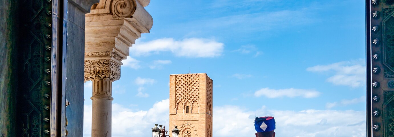 Una persona vestida con traje tradicional frente a un puerta ricamente decorada, con un cielo azul despejado y una torre distante.