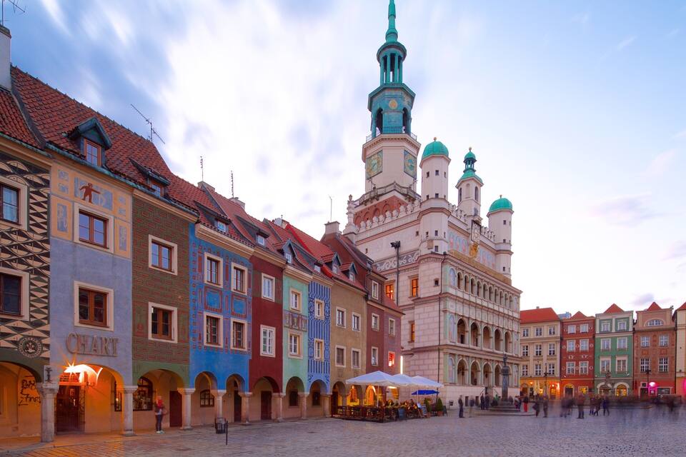 Uma praça histórica com edifícios coloridos, uma igreja imponente com torre do relógio e um calçadão de paralelepípedos.