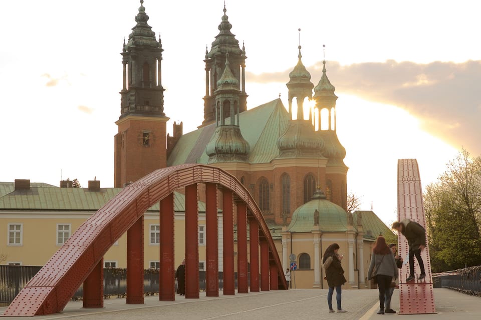 A bridge with red pillars and a large church with green domes in the background.