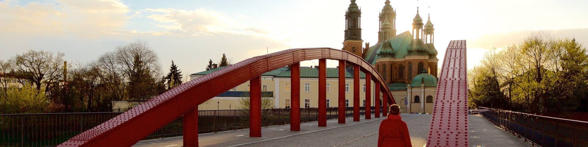 A pedestrian bridge with red supports and a tall red sculpture in a park with a historic building in the background.