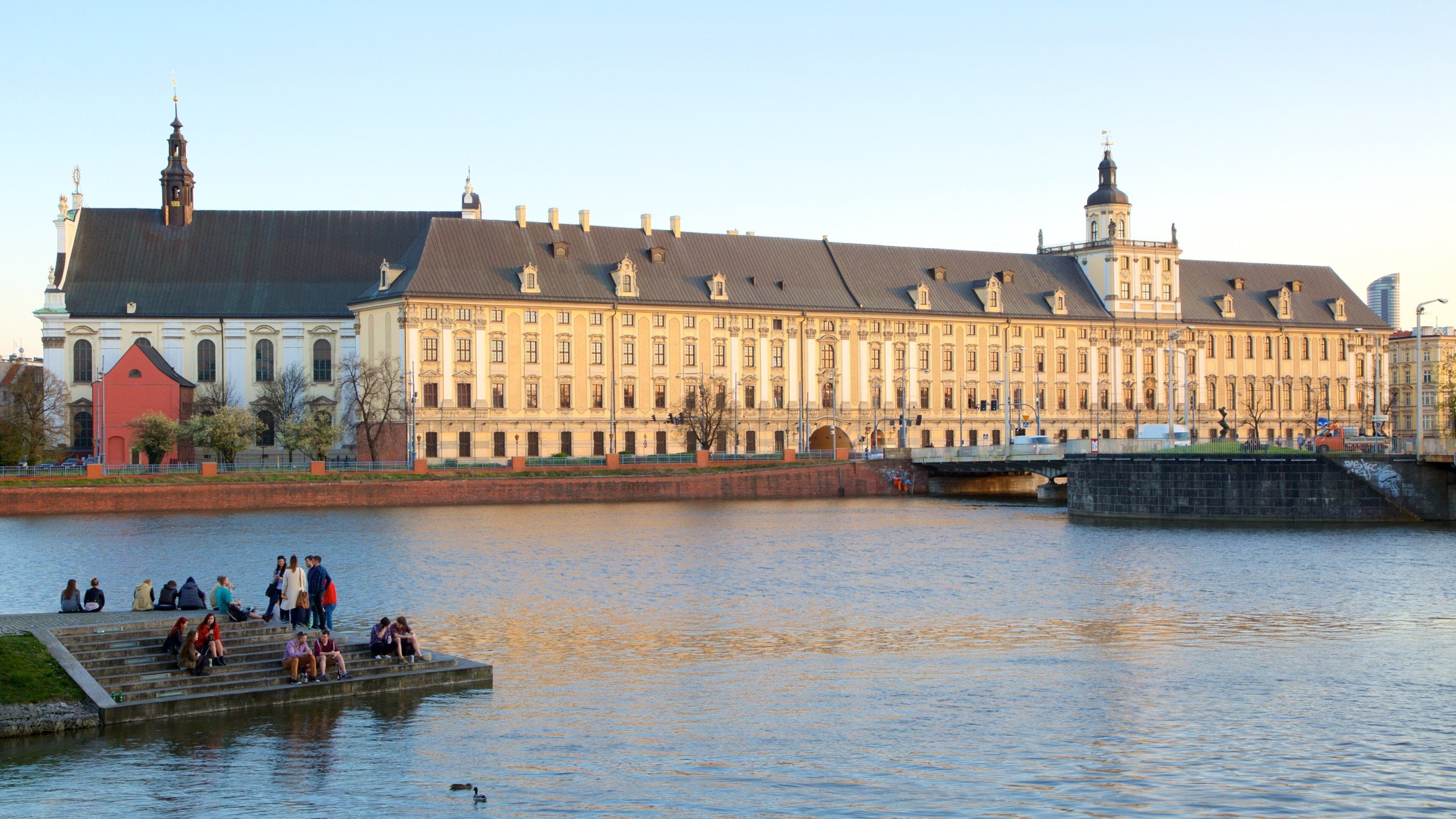 A historic building with a large central tower, a red-roofed structure to the left, and a bridge over a river.