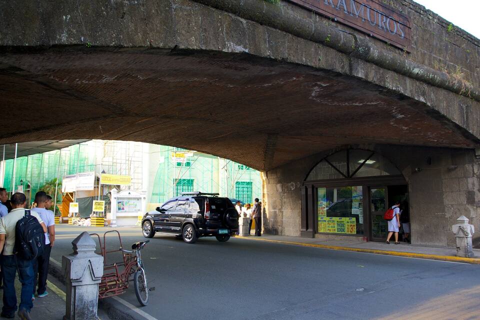 A busy street under a large stone bridge with people waiting and a black SUV driving by.
