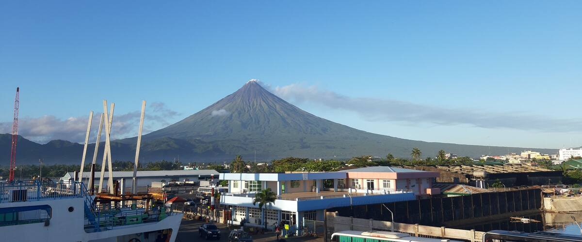 A coastal town with a prominent volcano in the background, a pier with boats, and a mix of commercial and residential buildings.