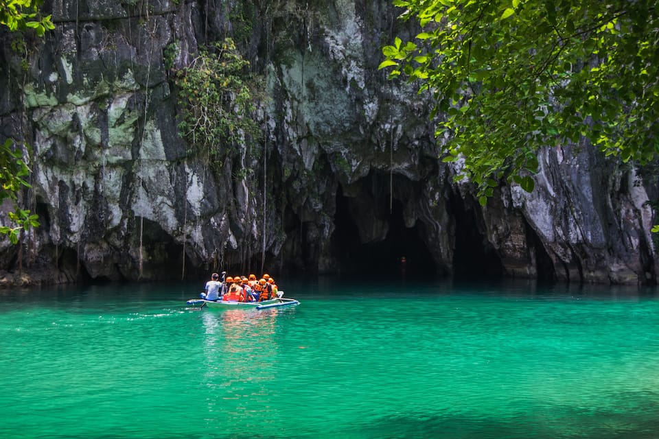 A group of people in a boat on turquoise water with a rocky cave wall in the background.