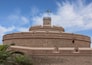 Una fortaleza de piedra histórica con una bandera en la parte superior, palmeras en primer plano y un cielo azul despejado.
