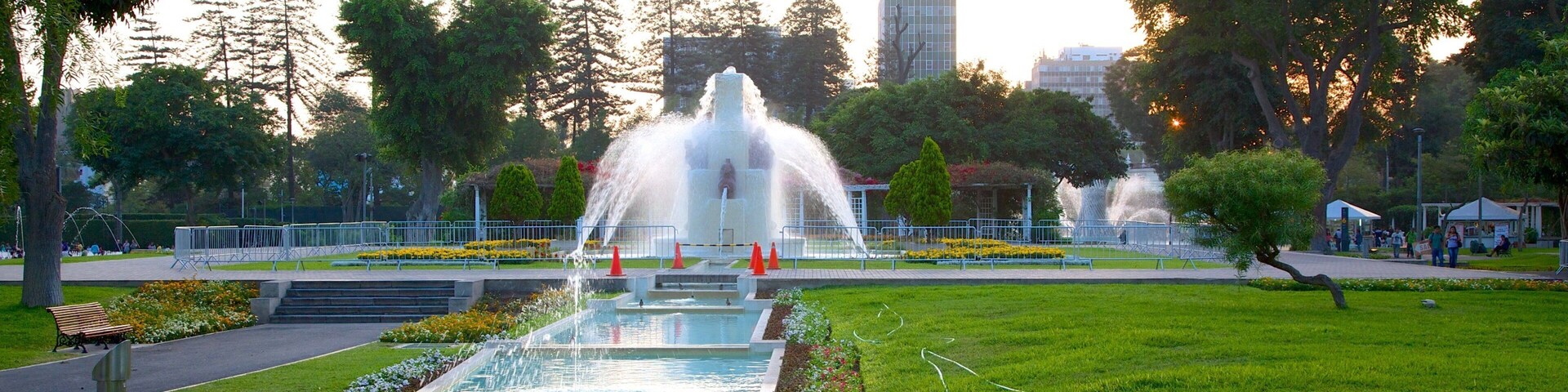 Un parque con una fuente, césped verde, árboles y un edificio al fondo.