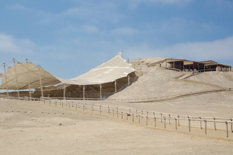 A large, open-air structure with a white canopy, surrounded by a sandy landscape and a staircase leading up to a building.