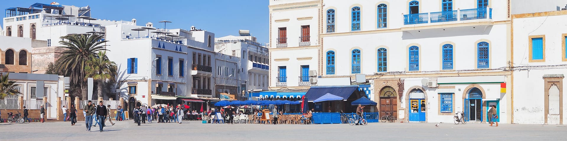 A bustling square with white buildings featuring blue doors and windows, palm trees, and a clear blue sky.