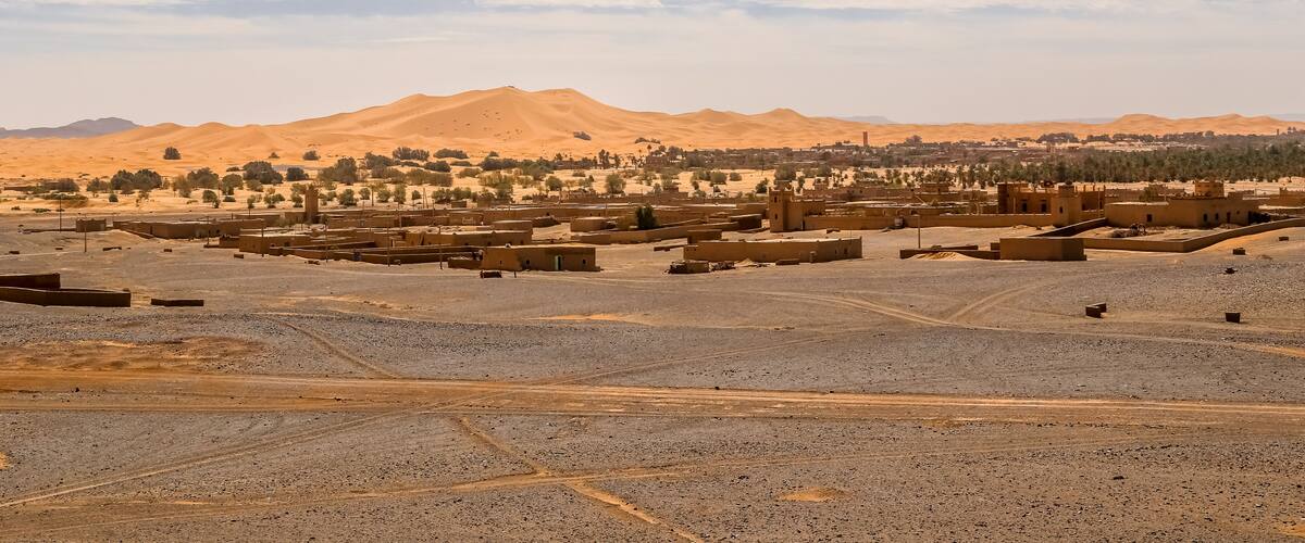 A desert landscape with sand dunes, a town with traditional architecture, and a mountain range in the background.