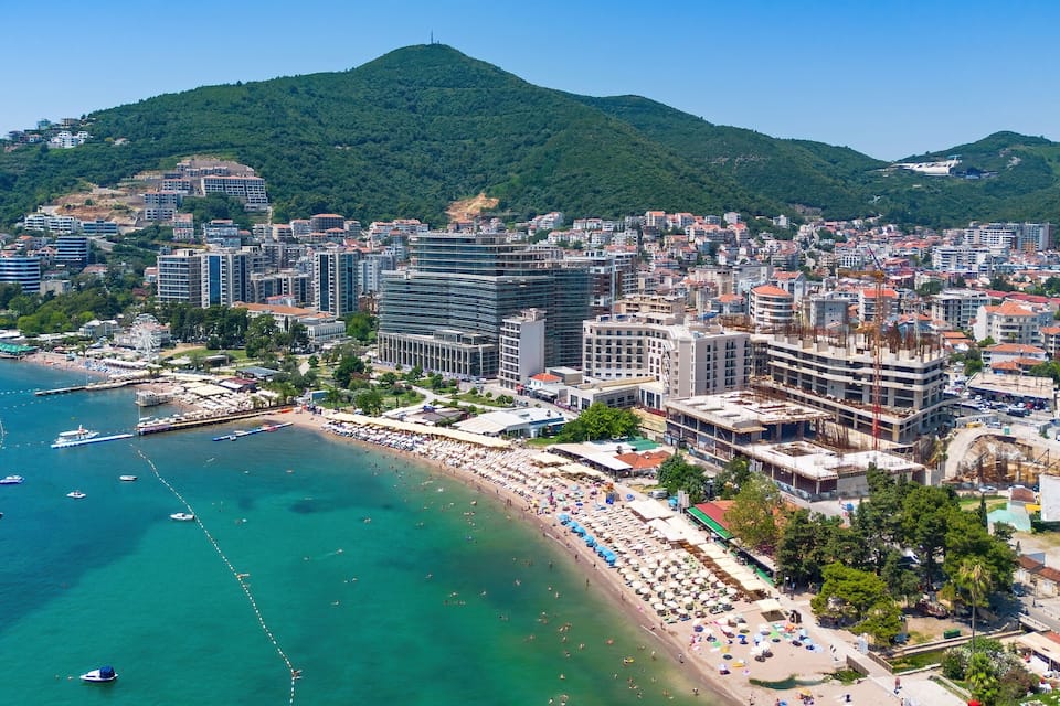 A coastal city with a beach, boats, and a mountainous backdrop.