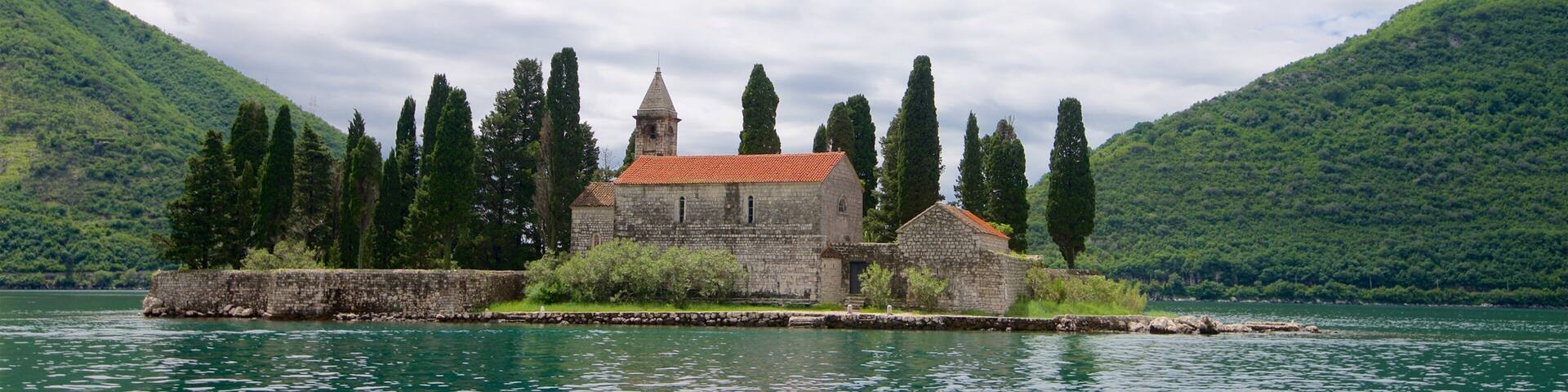 A stone church with a red roof by a calm lake, surrounded by green hills and tall cypress trees.