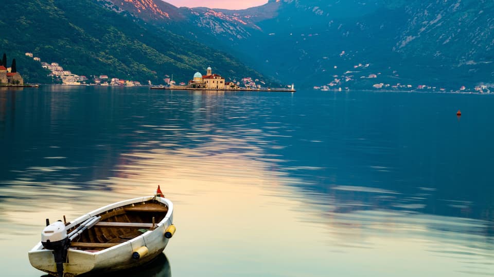 A calm lake with a wooden boat anchored near the shore, surrounded by mountains and a small village with buildings.