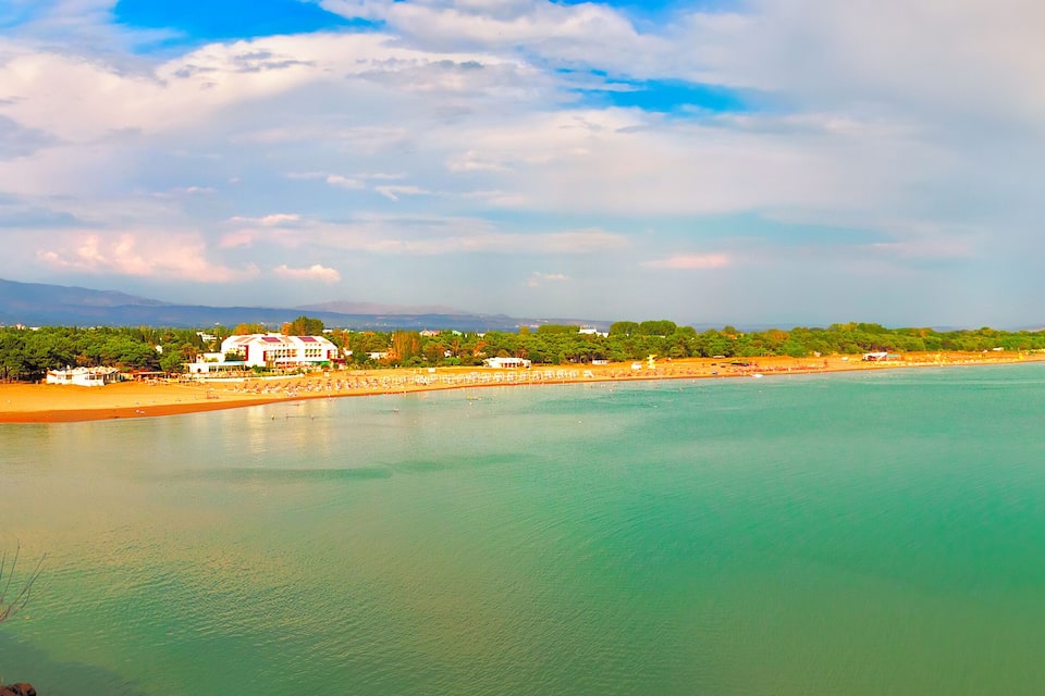 Een kustlandschap met een strand, helder turquoise water en een achtergrond van bergen en een gedeeltelijk bewolkte lucht.