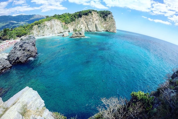 A coastal landscape with clear turquoise waters, rocky cliffs, and a distant mountain range.