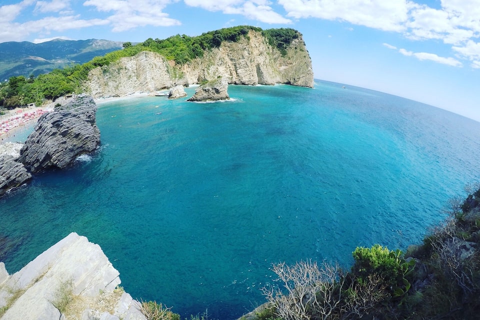 A coastal landscape with clear turquoise waters, rocky cliffs, and a distant mountain range.