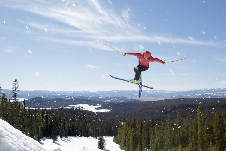 A skier mid-air performing a jump over a snowy mountain slope with a forested valley in the background.