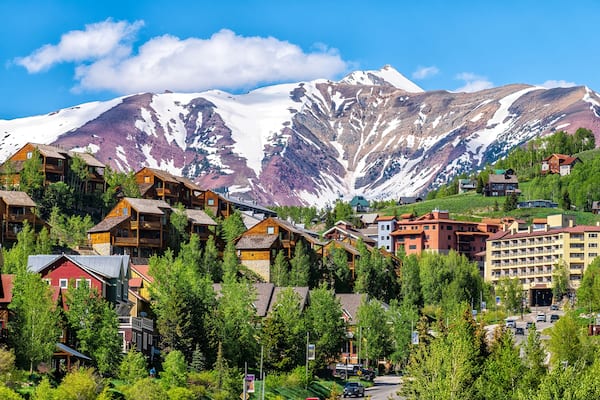 Mount Crested Butte ski resort town, Colorado with houses homes, wooden lodge hotels on hill in summer and green trees by snow-capped mountains in background