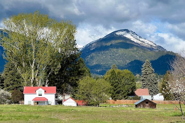 A rural landscape with a white house with a red roof, a wooden barn, and a large mountain with snow-capped peaks in the background.