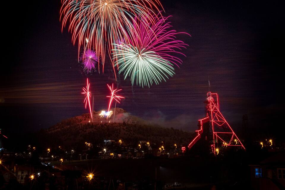 A night scene with fireworks over a town, a hill with houses, and a large red-lit structure.