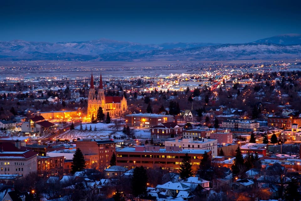 A cityscape at dusk with illuminated buildings and a prominent cathedral with two spires. Snow-covered mountains are visible in the background.