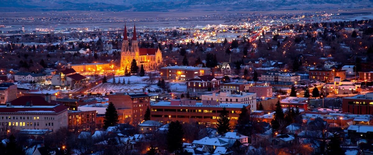 A cityscape at dusk with illuminated buildings and a prominent cathedral with two spires. Snow-covered mountains are visible in the background.