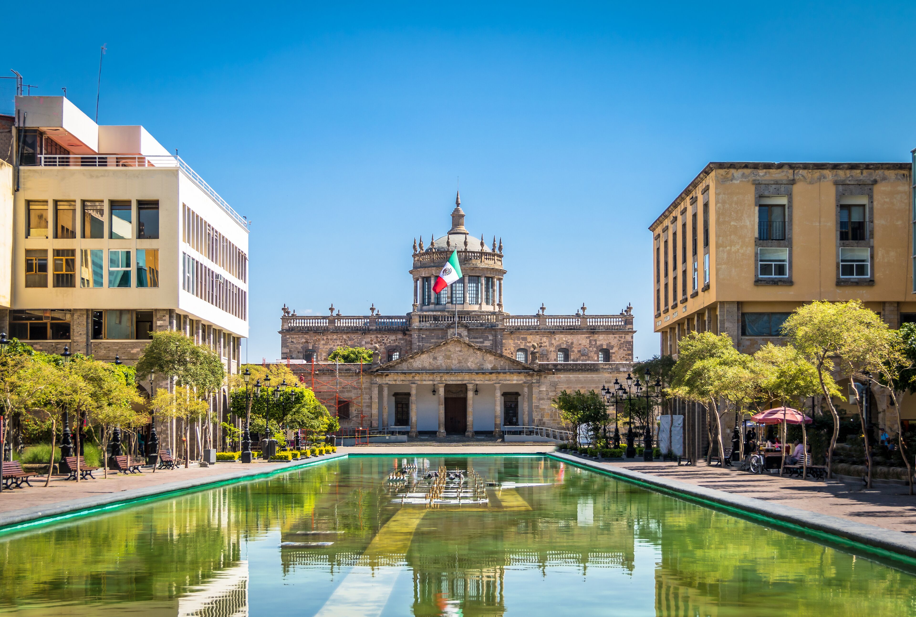 A historic building with a central dome, a flag, and a reflective pool in front.
