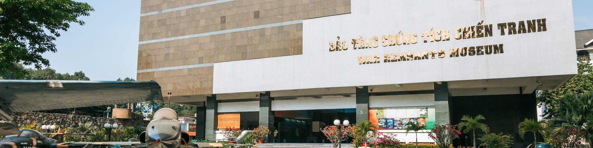 A museum building with a large sign in a foreign language, surrounded by potted plants and a display of military aircraft.