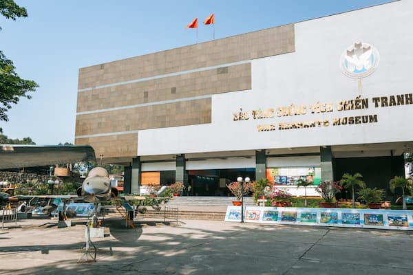 A museum building with a large sign in a foreign language, surrounded by potted plants and a display of military aircraft.