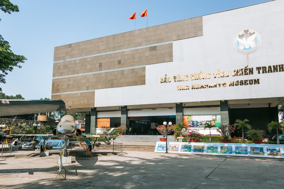 A museum building with a large sign in a foreign language, surrounded by potted plants and a display of military aircraft.