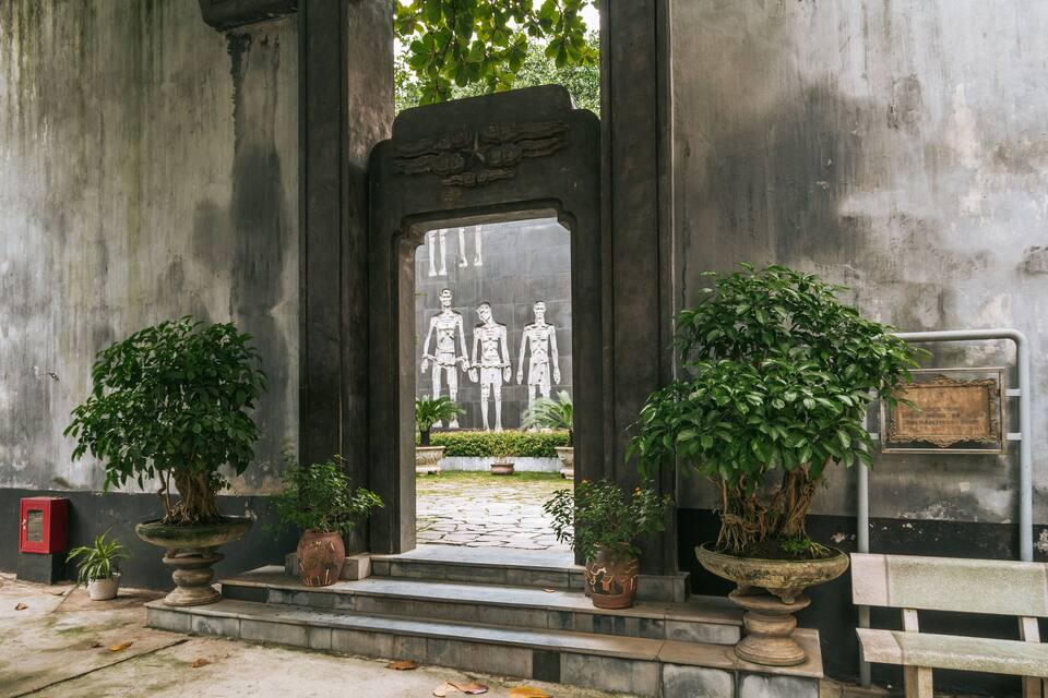 A stone entrance with a relief sculpture of three figures, flanked by potted plants and a bench.