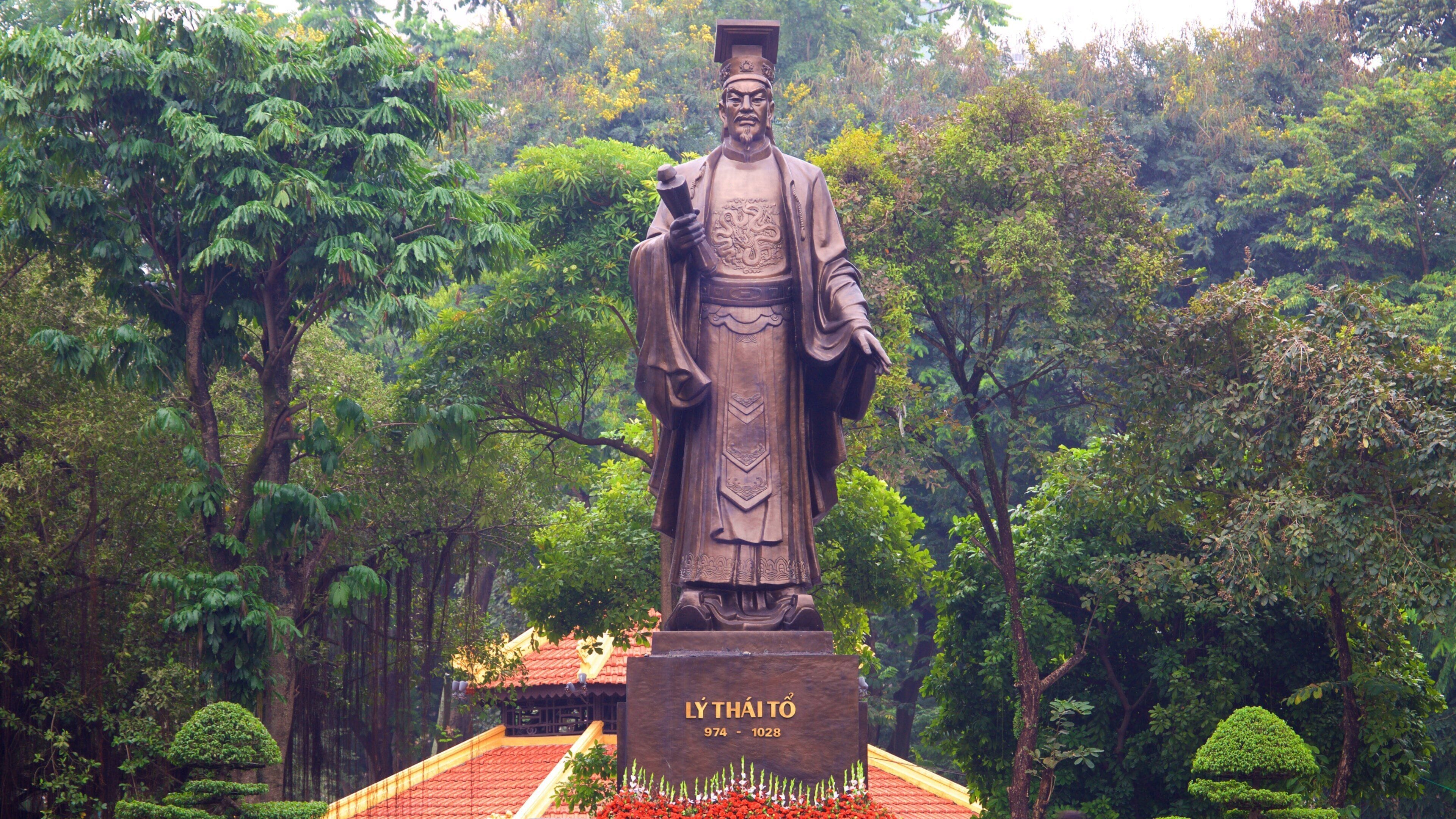 A statue of a historical figure in a lush garden with a red-roofed pavilion.