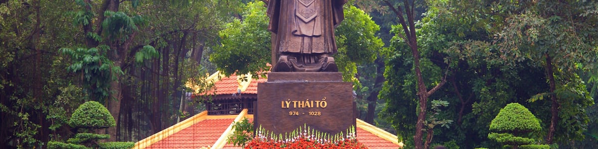 Une statue d’une figure historique, dans un jardin luxuriant, avec un pavillon au toit rouge.