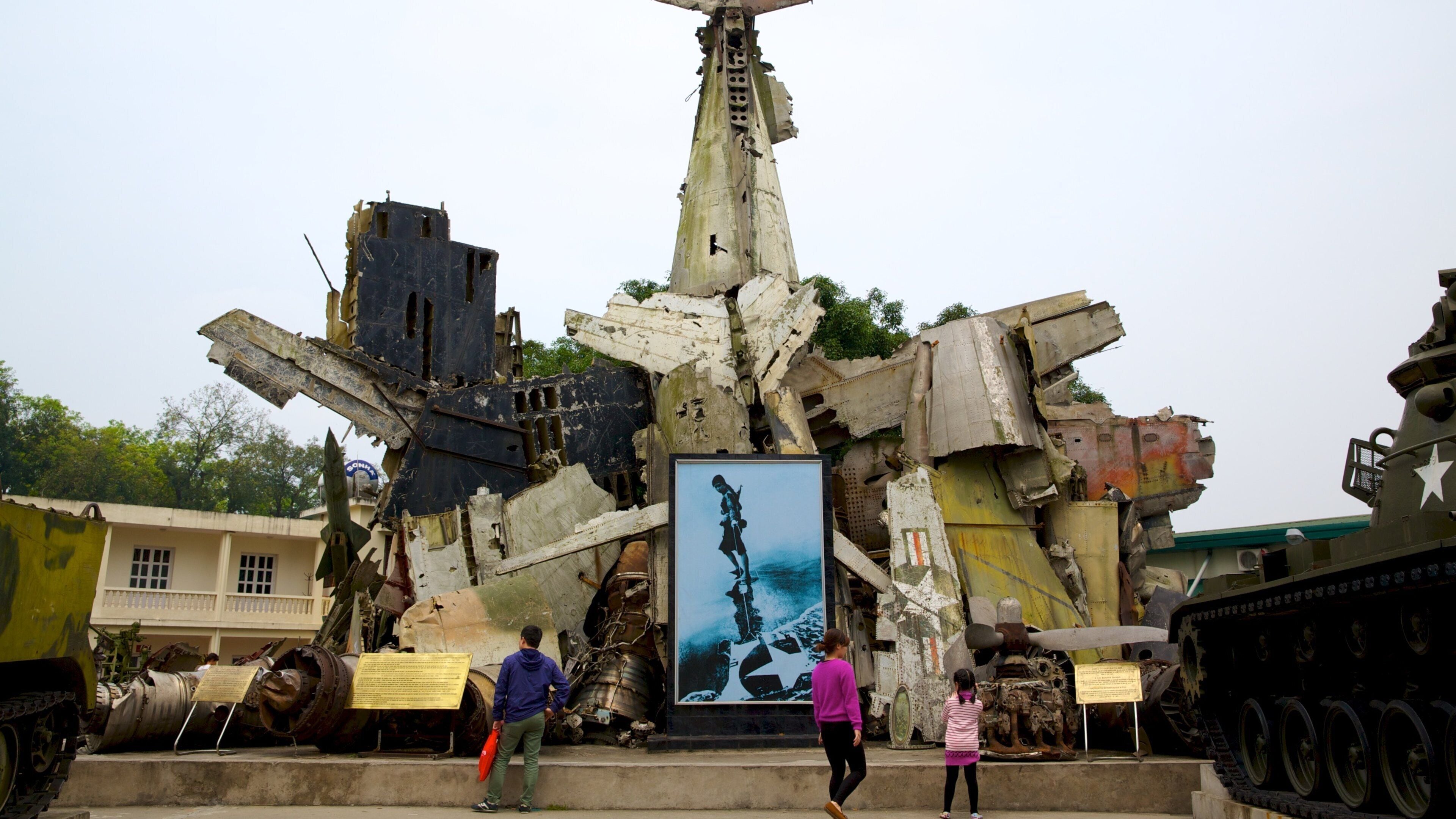 Uma aeronave militar grande e enferrujada com insígnia de estrela, rodeada por outros veículos militares e um monumento com o perfil de um soldado.
