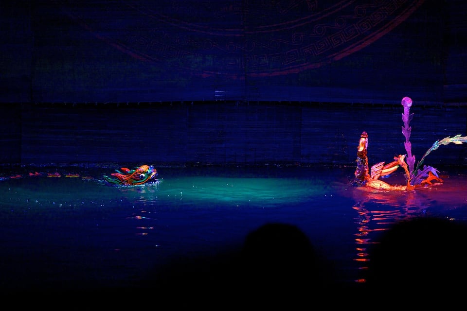 A stage with performers in costumes, a water feature, and a dark background.