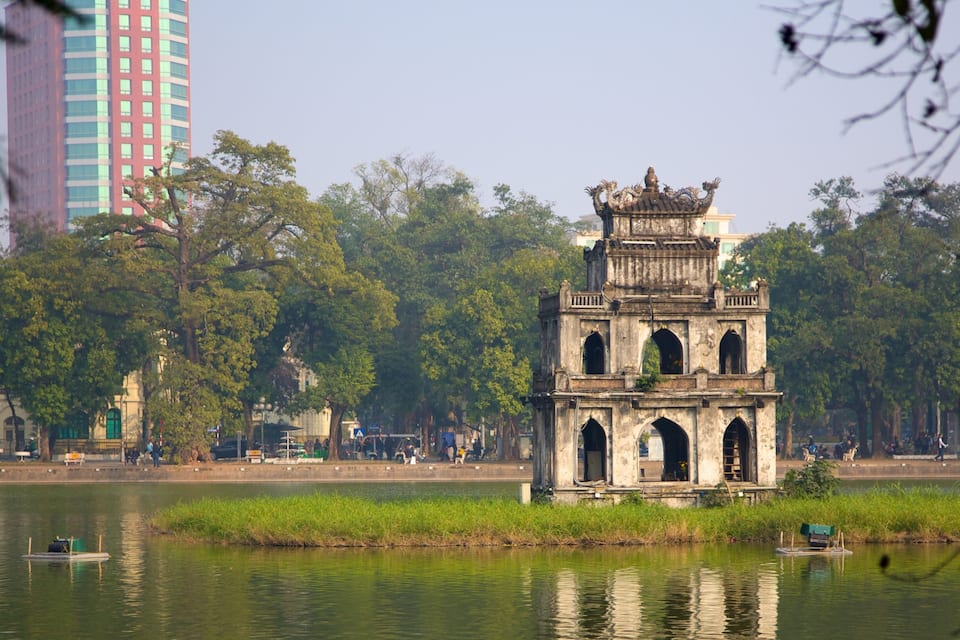 Ein historischer Turm an einem See, umgeben von Bäumen und modernen Gebäuden.