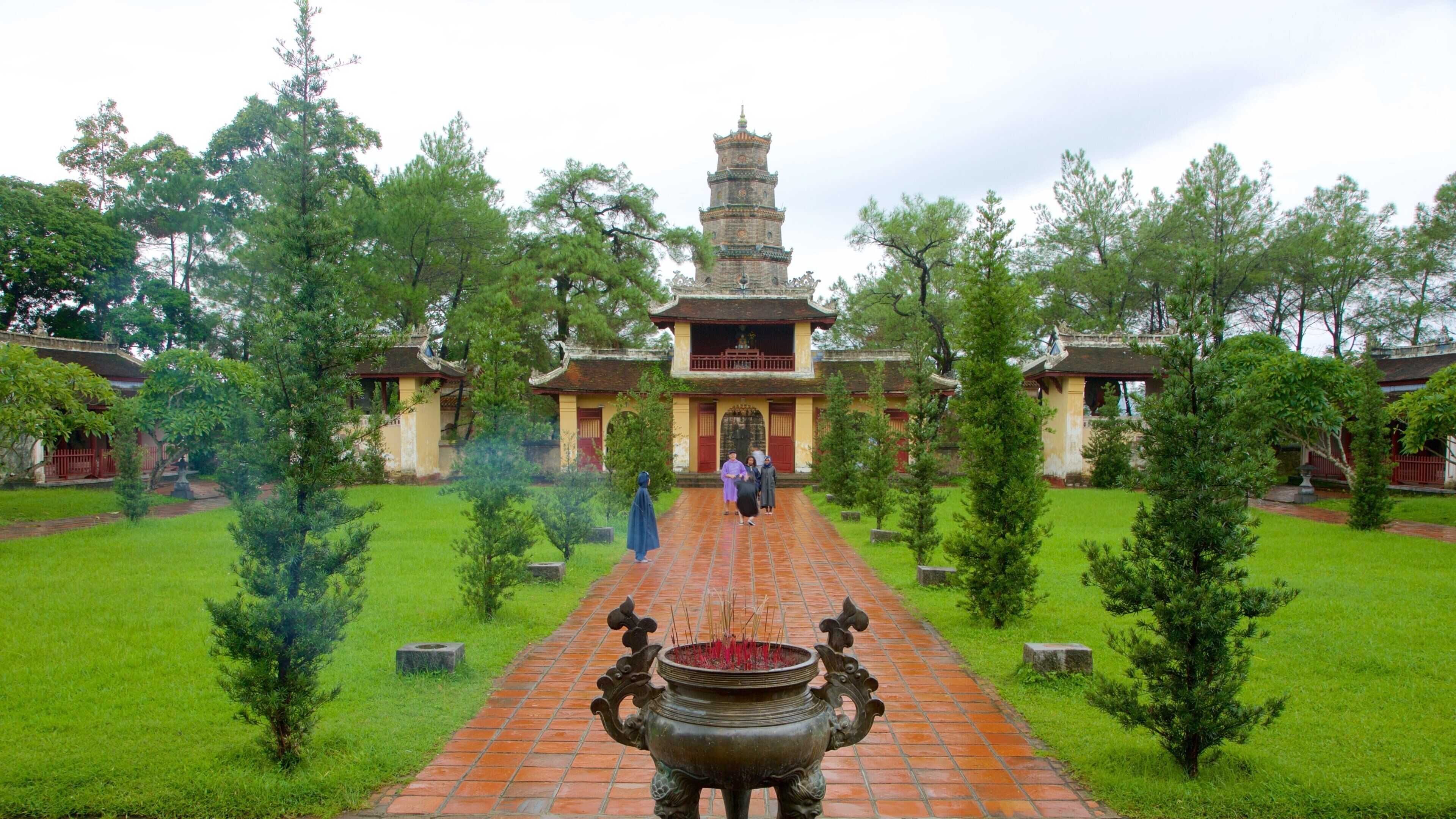 A traditional temple with a central tower, surrounded by a garden with small trees and a brick pathway leading to a large incense burner.
