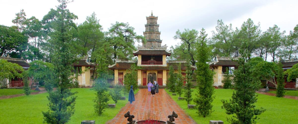 A traditional temple with a central tower, surrounded by a garden with small trees and a brick pathway leading to a large incense burner.