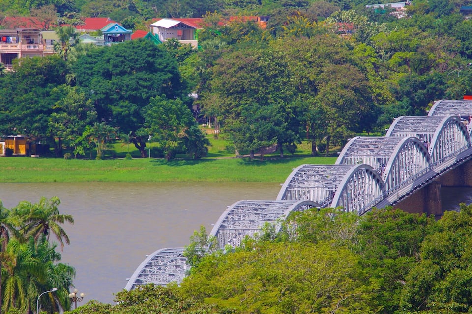 Un puente sobre un río con vegetación exuberante y edificios al fondo.